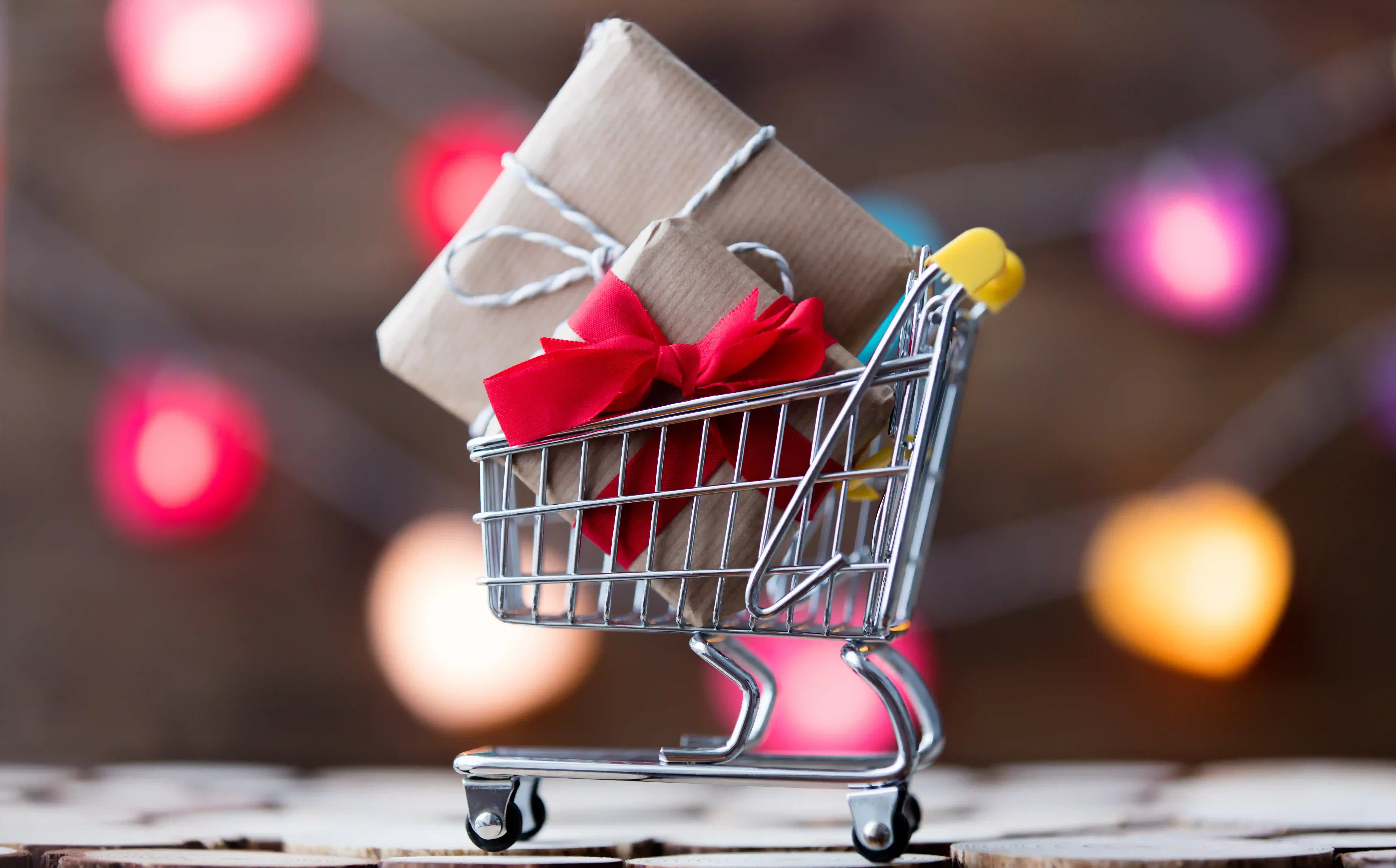 AdobeStock_129302573 Shopping cart with gifts inside sitting in front of faded holiday. string-lights